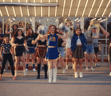 A group of people, including a person in a cheerleader outfit, dance energetically in front of a building with striped lights above them