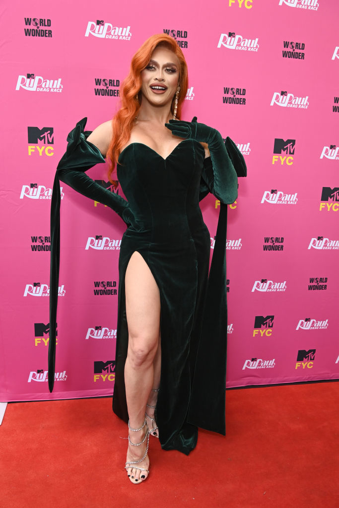 Person posing on a red carpet, event backdrop behind them, wearing a strapless gown with high slit and long gloves
