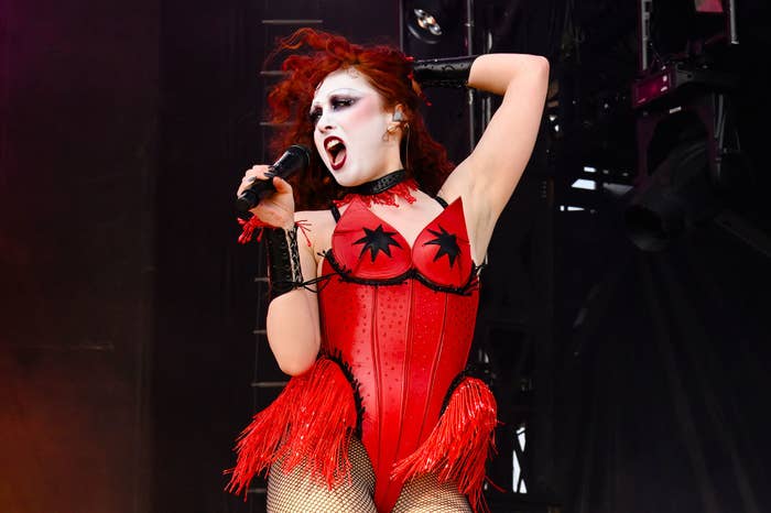 Performer in a dramatic red outfit, including a corset and fringed details, sings passionately into a microphone on stage