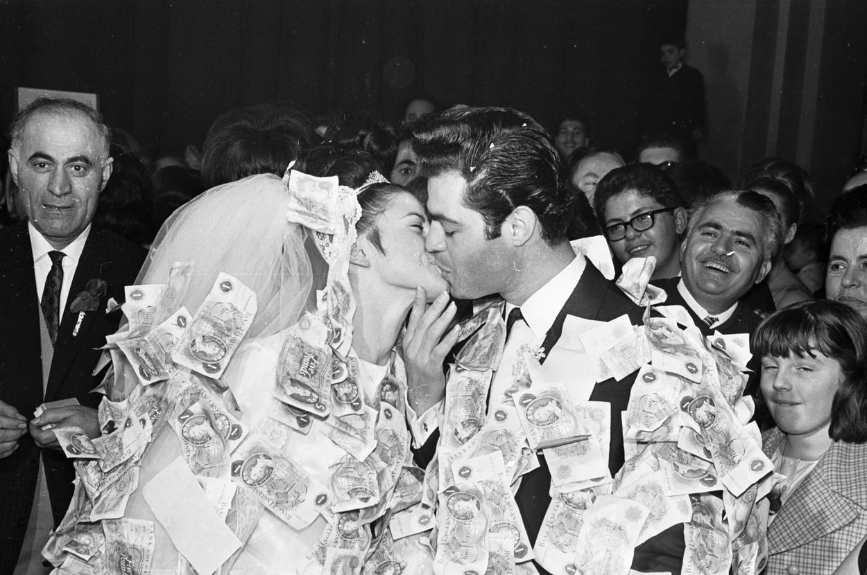 Bride and groom kiss on their wedding day, their outfits covered in paper money. Guests, including an older man and young girl, smile and watch