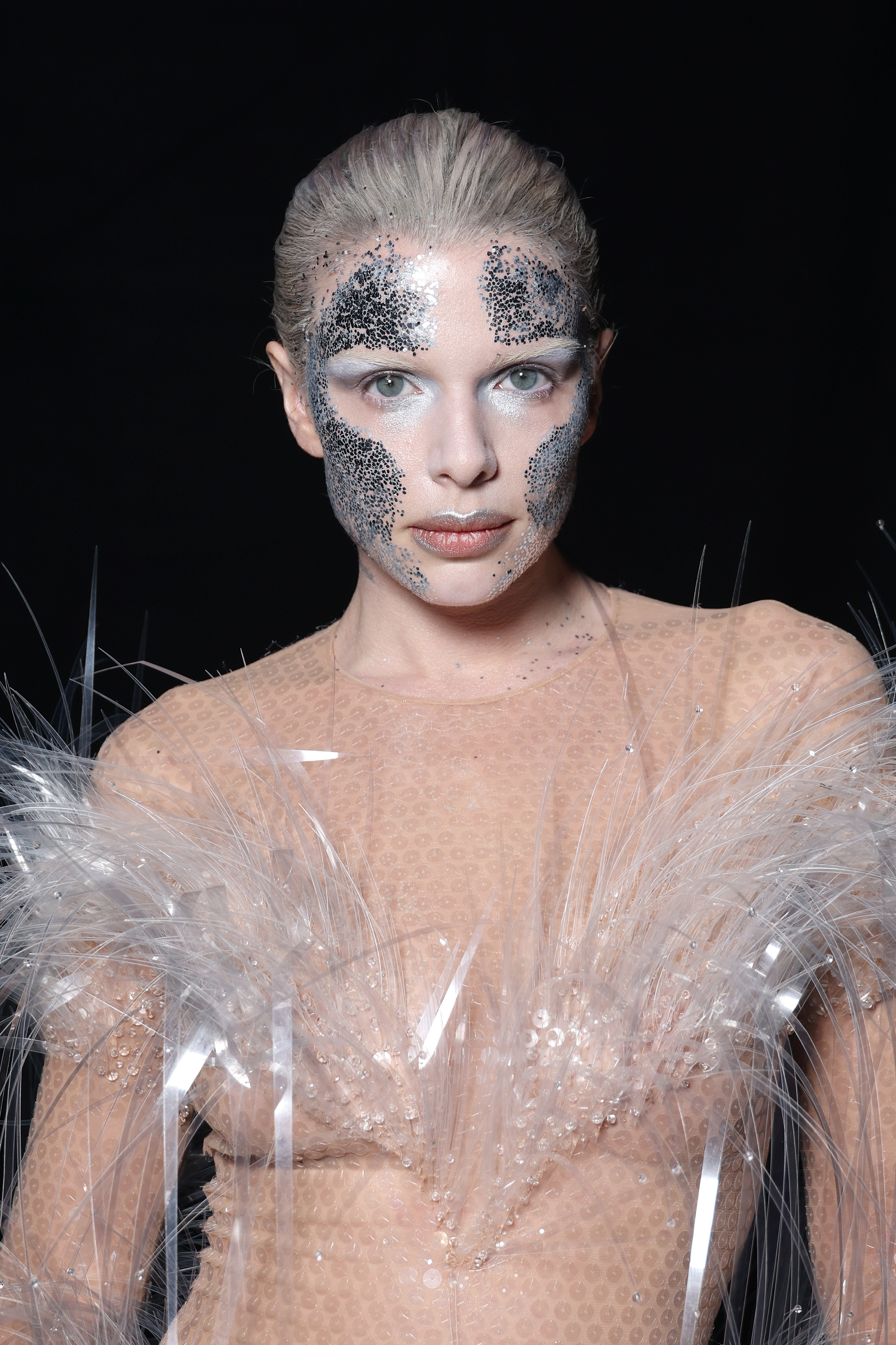 Julia Fox wearing a sheer, textured outfit with feather detailing and glitter makeup on face, posing against a black background
