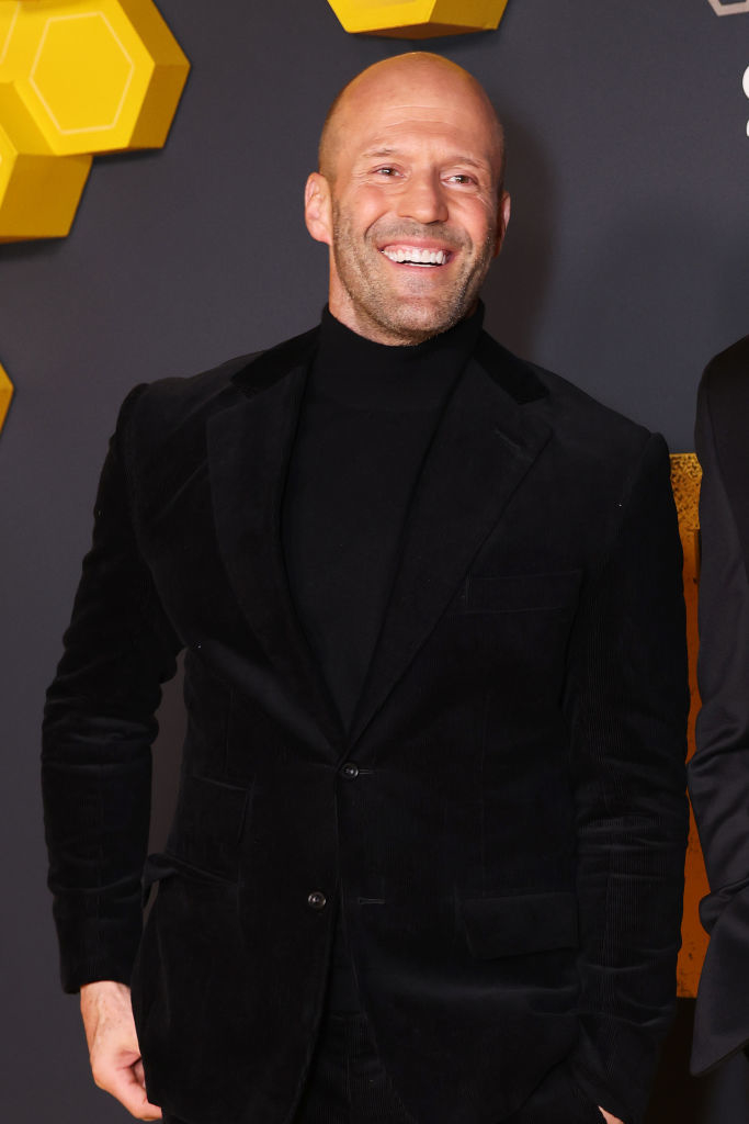 Jason Statham smiling on a red carpet, wearing a black turtleneck sweater and black suit
