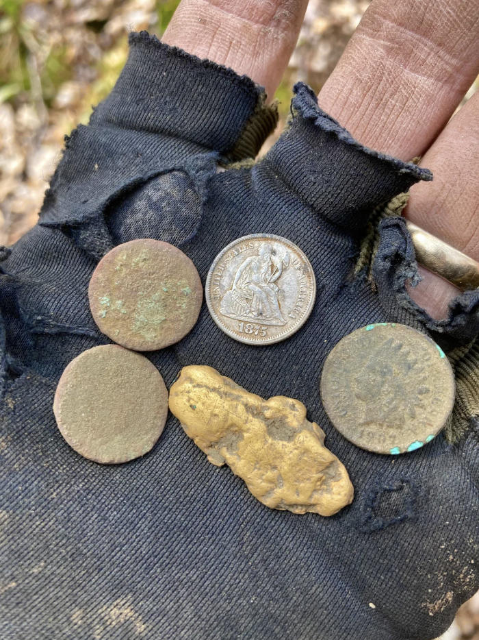 Person wearing a black, tattered glove is holding four old coins and a gold nugget in their hand. Coins show wear and patina