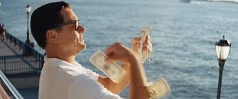 Leonardo DiCaprio, wearing sunglasses and a white shirt, stands by the water, throwing money into the air