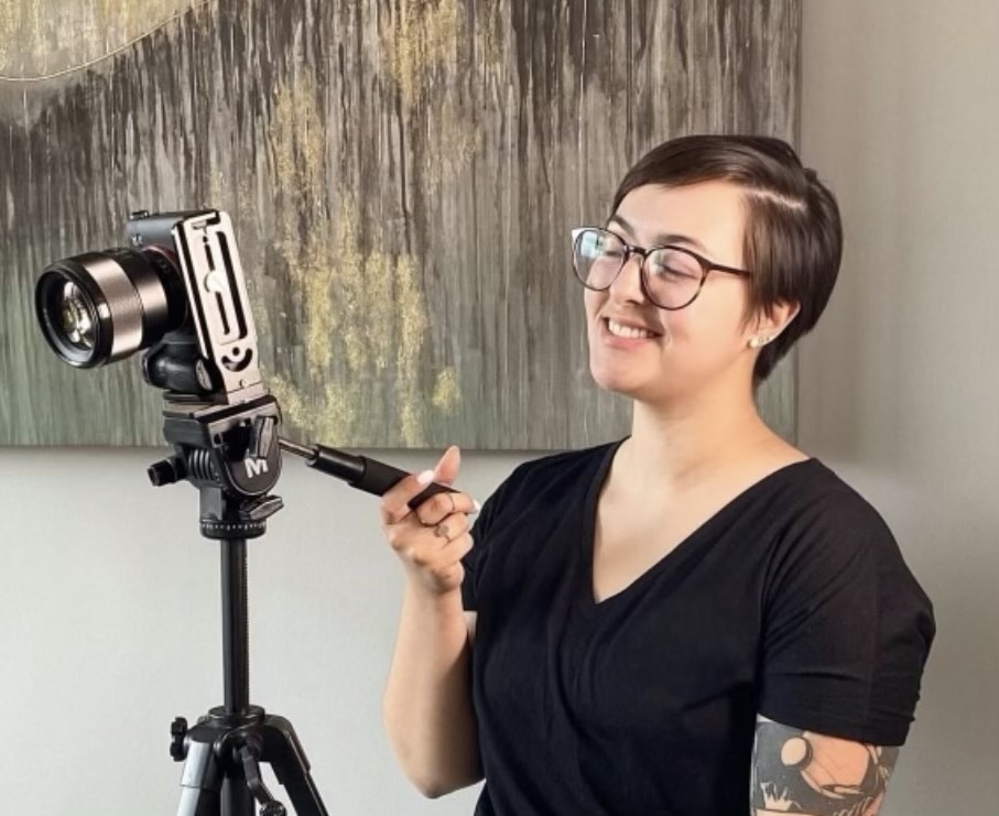Teresa, with short hair and glasses, wearing a dark shirt, smiling and setting up a camera on a tripod
