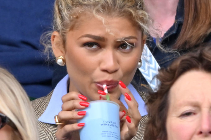 Zendaya drinks from a cup using a straw, casually dressed in a blazer, with a crowd in the background