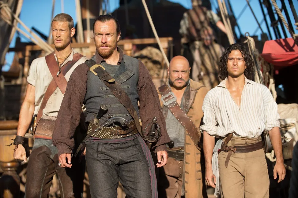 Cuatro personajes de la serie "Black Sails" caminan por un barco: Toby Stephens, Tom Hopper, Mark Ryan y Luke Arnold, vestidos con ropa de época pirata