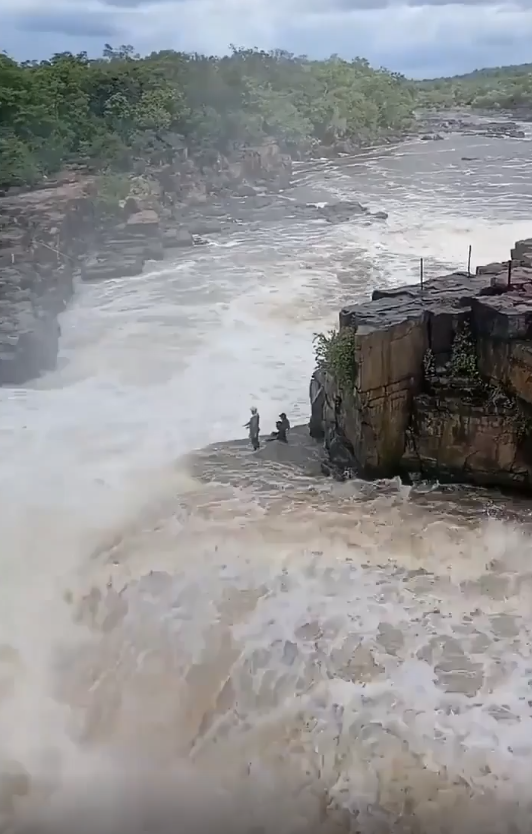 Three people stand dangerously close to the edge of a large, powerful waterfall. Dense forest surrounds the rushing water