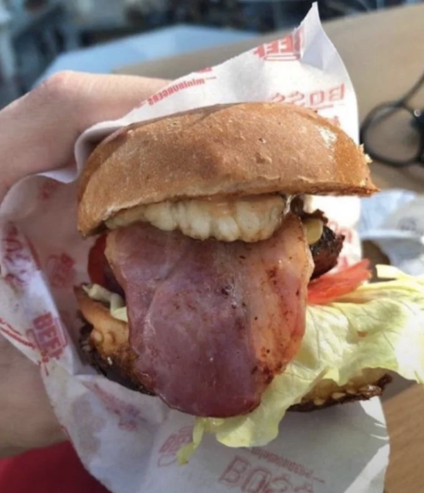 A hand holds a burger with bacon, lettuce, tomatoes, and an unusual topping: what appears to be an artificial denture set, creating a humorous effect