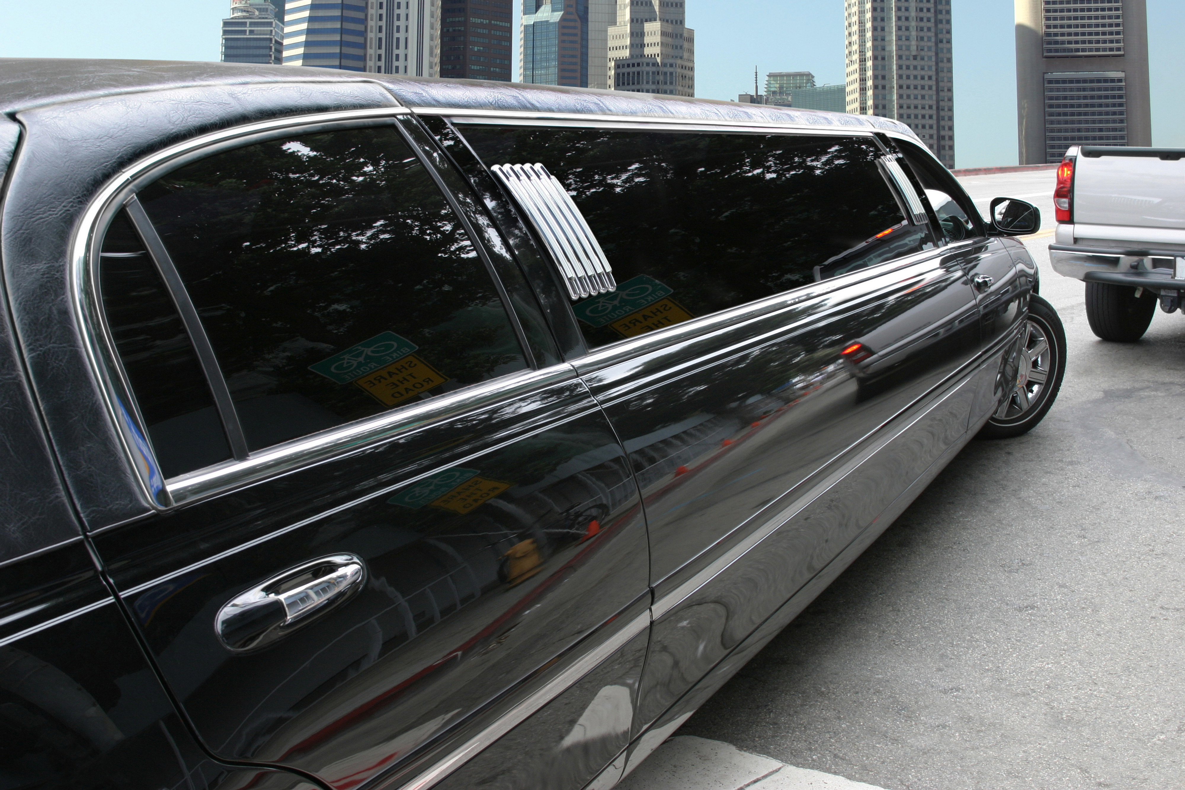 A long black limousine is parked on a city street near a white truck, with tall buildings visible in the background
