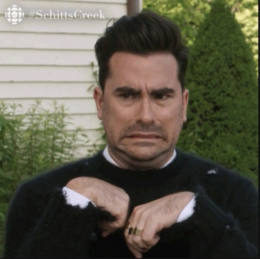Dan Levy making a funny expression with hands held up near his chest in a scene from Schitt’s Creek. #SchittsCreek visible in top corner