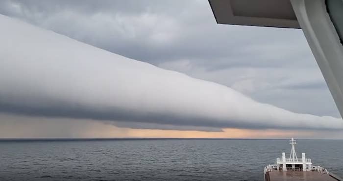 A ship sails towards a unique, dense, long horizontal cloud formation stretching across the sky above the ocean