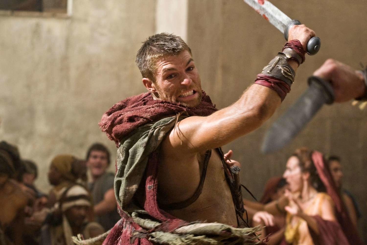 Hombre musculoso blandiendo una espada en medio de una batalla en lo que parece ser un antiguo escenario romano. Otros combatientes se ven al fondo