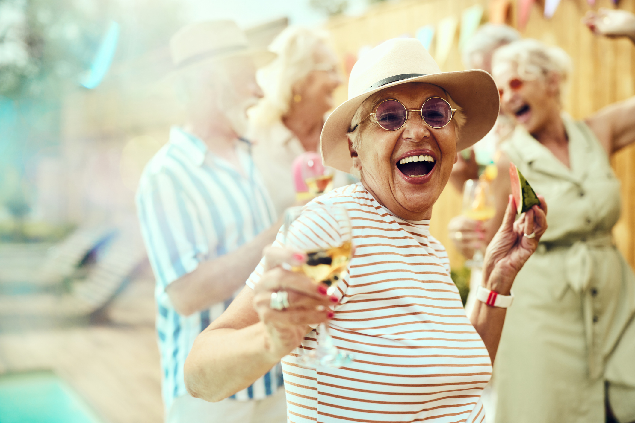 Elderly individuals enjoy a joyful outdoor gathering, smiling and holding drinks. Prominent in the image, a woman wearing a hat and glasses, laughs with a drink in hand