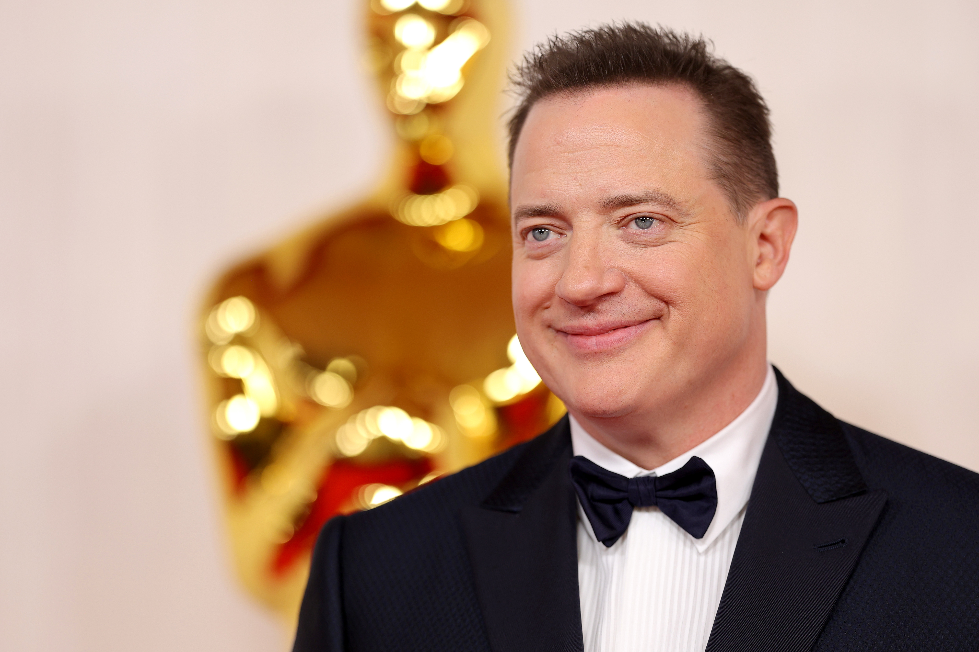 Brendan Fraser at a red carpet event, dressed in a tuxedo with a bow tie, smiling. An Oscar statue is blurred in the background