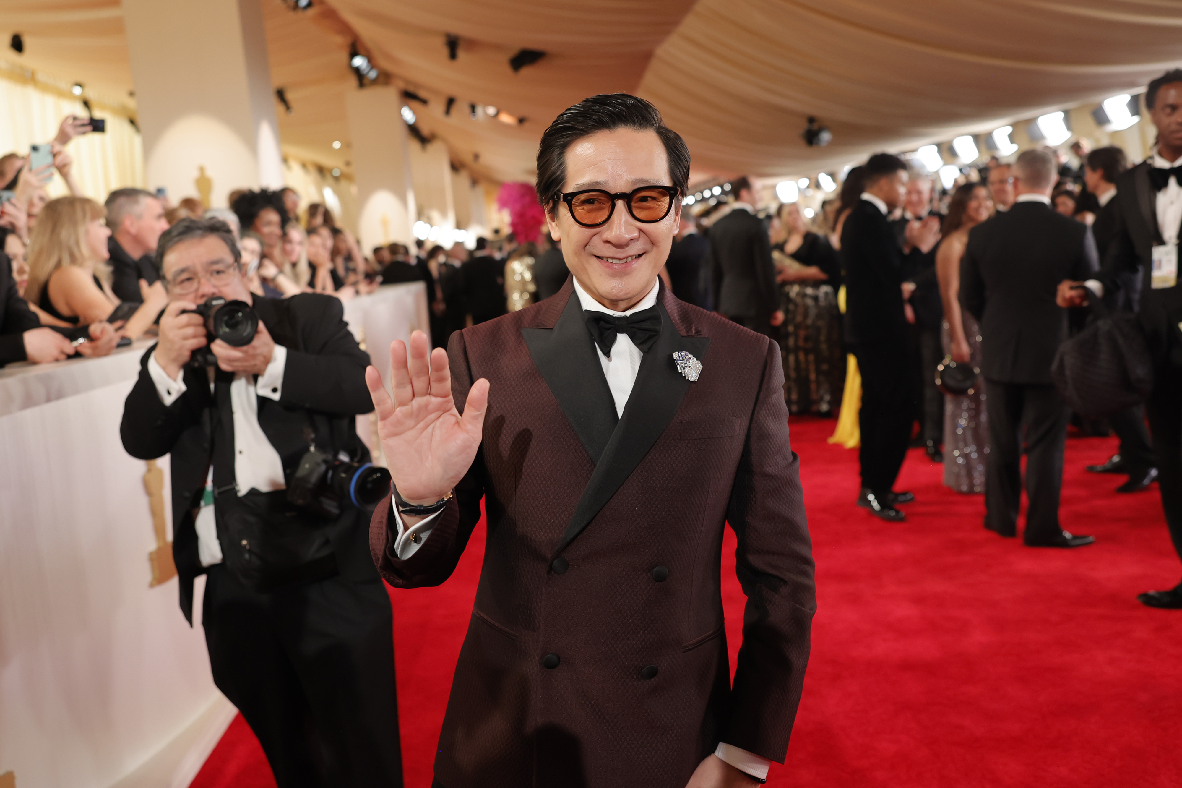 Ke Huy Quan waves on the red carpet, wearing a double-breasted suit, surrounded by photographers and guests