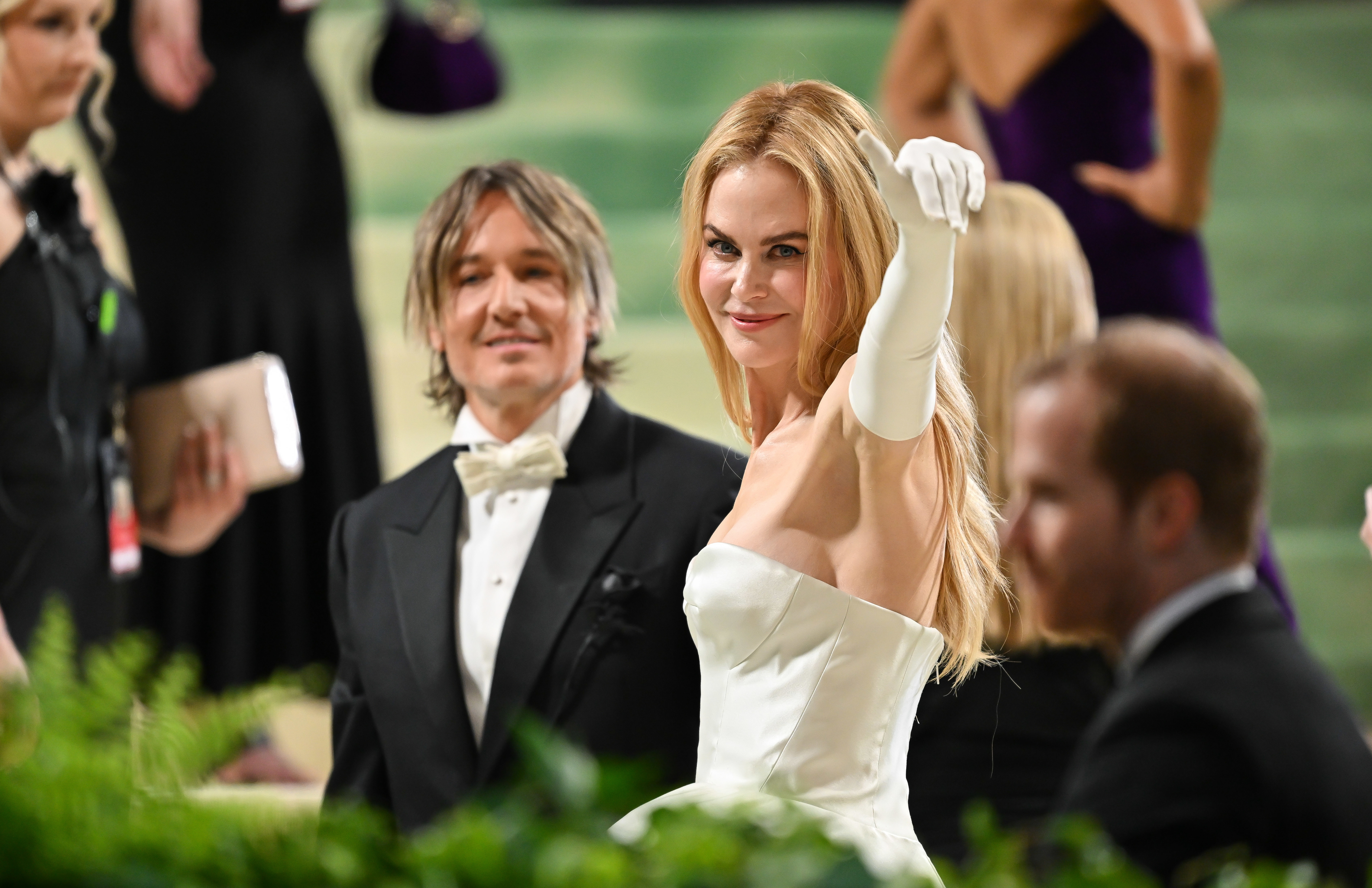 Nicole Kidman in a strapless gown, waving, with Keith Urban in a tuxedo at a formal event. Other attendees are in the background