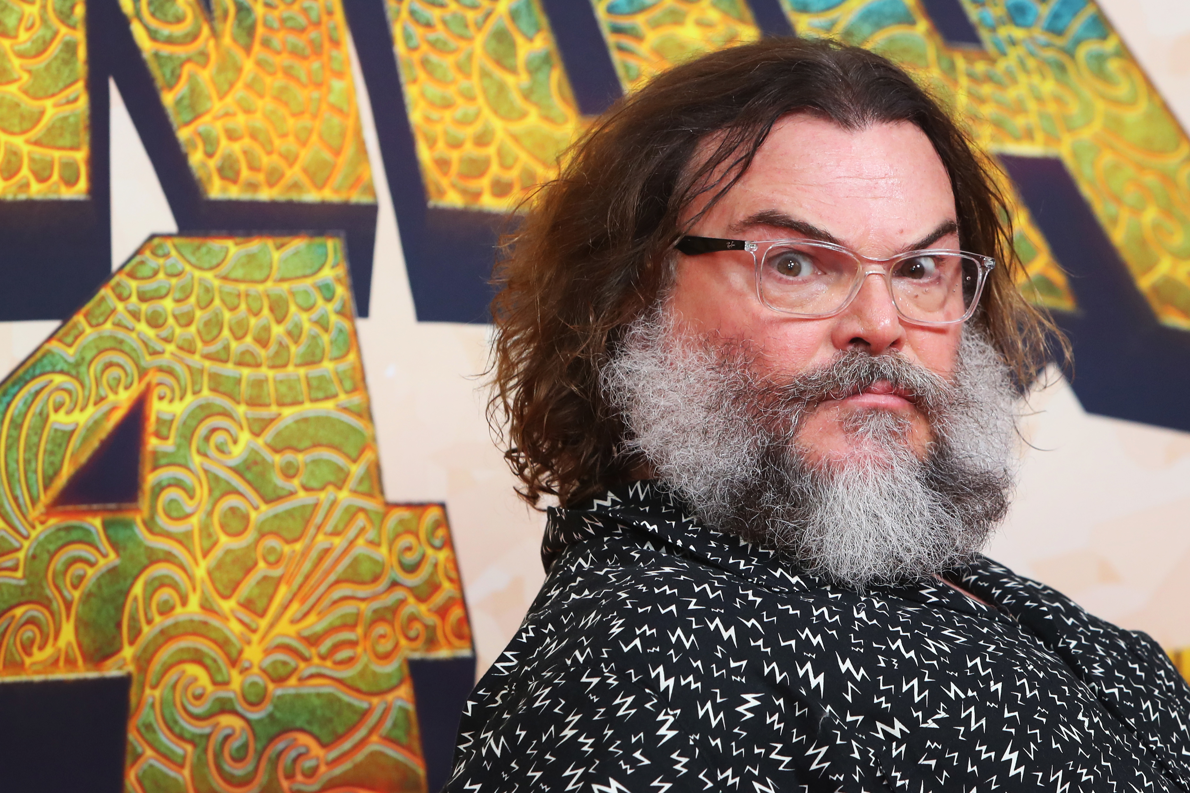 Jack Black with a thick beard and glasses poses in a patterned shirt at a movie premiere backdrop