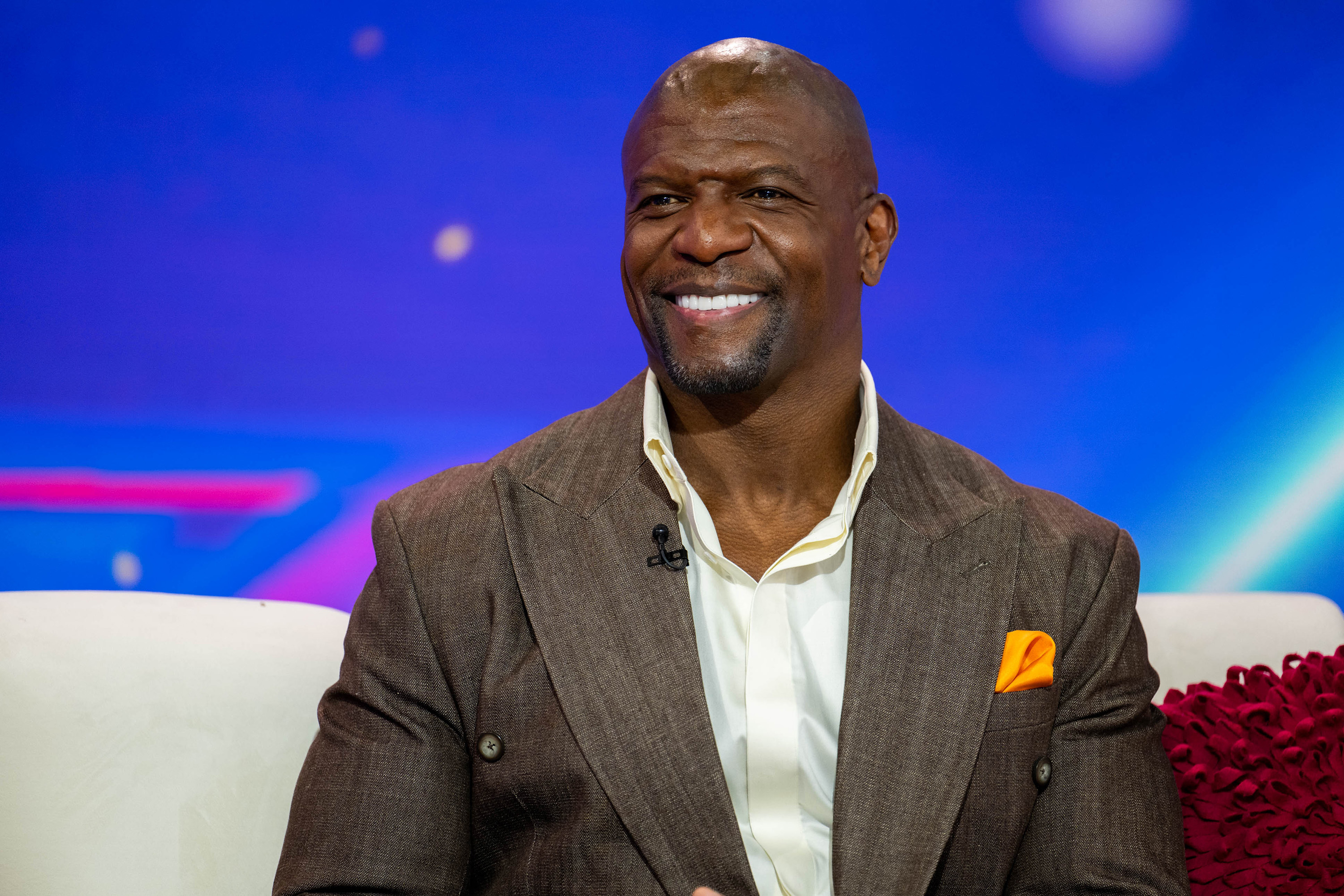Terry Crews, smiling while seated on a white couch, wearing a brown blazer, white shirt, and orange pocket square