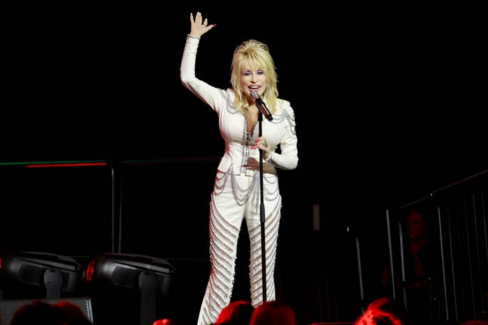 Dolly Parton waves while performing on stage, wearing a sparkling white jumpsuit with elaborate patterns and rhinestone accents, holding a microphone