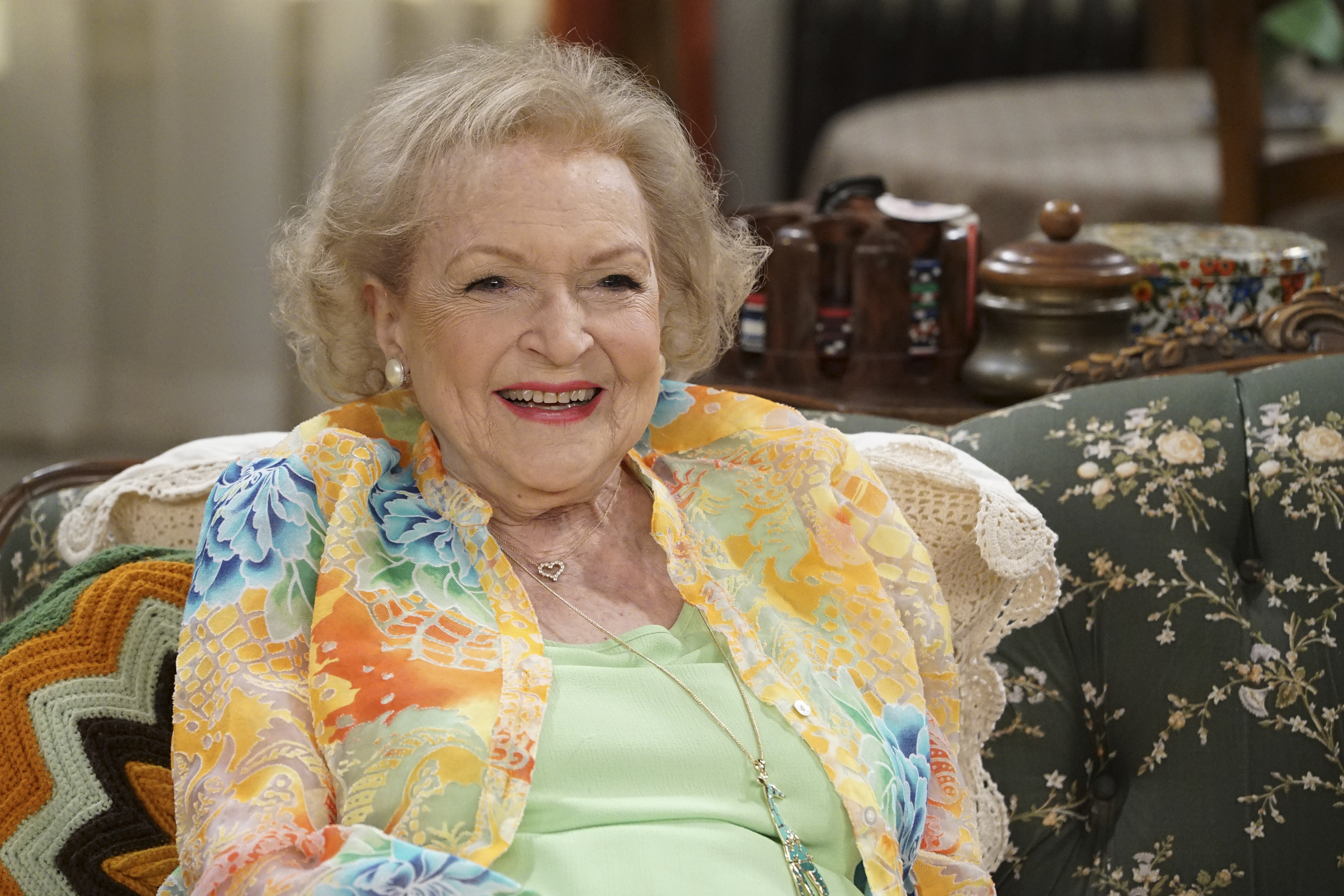 Betty White smiling, seated on a floral-patterned couch, wearing a colorful top over a light green blouse