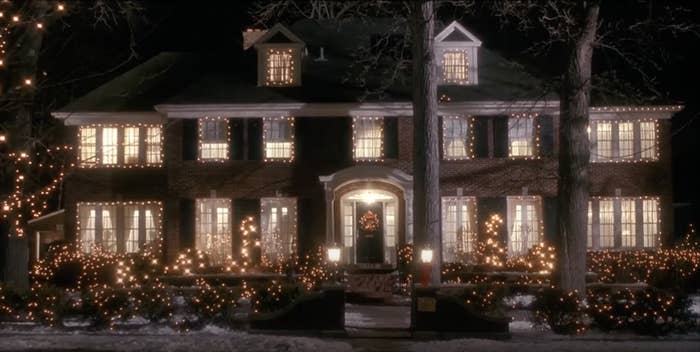 A large house decorated with numerous holiday lights shines brightly at night amidst trees and a light dusting of snow