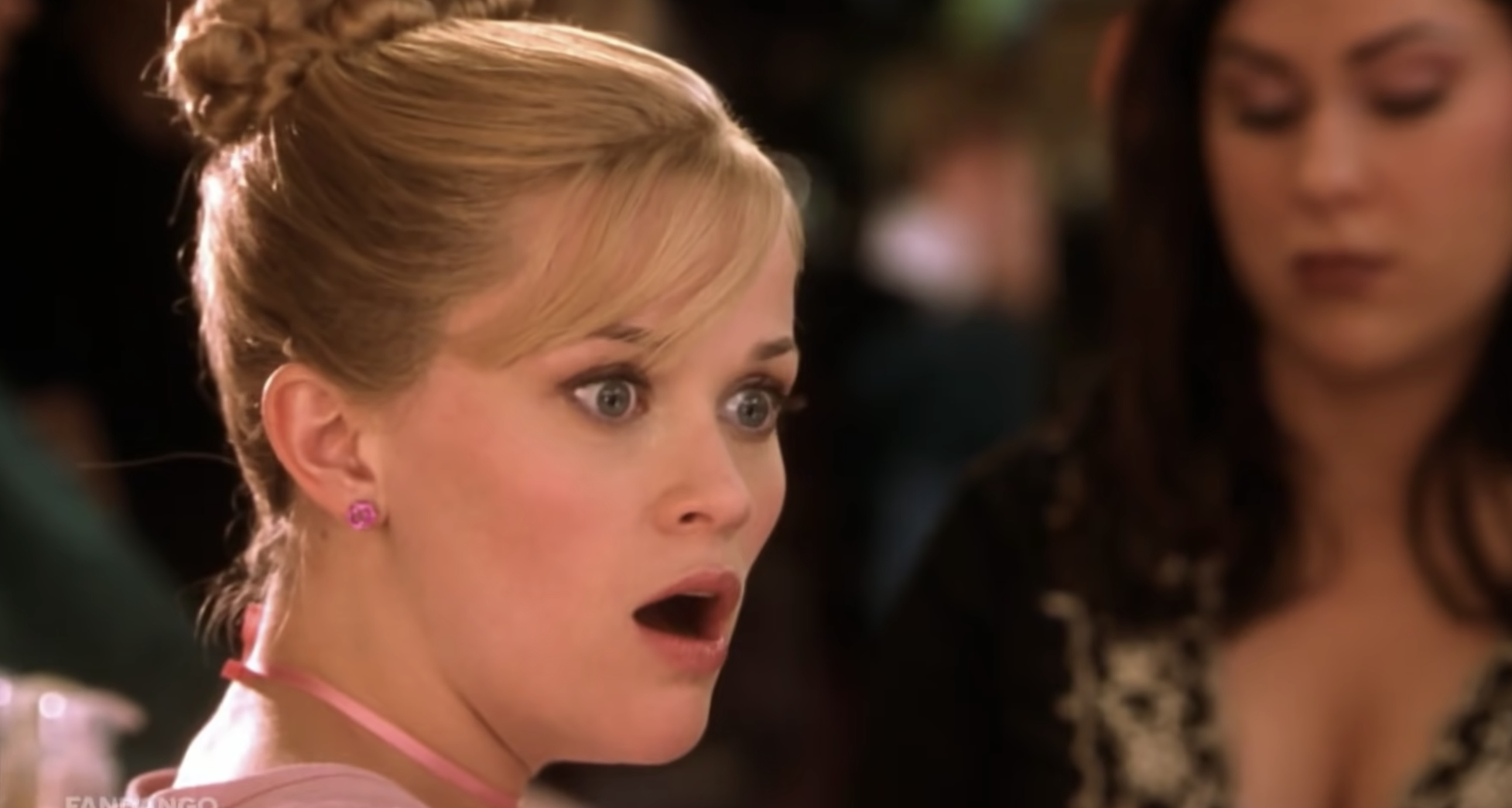 Reese Witherspoon, with a surprised expression, wearing a pink top and updo hairstyle, in a scene from a movie. Another woman is in the background