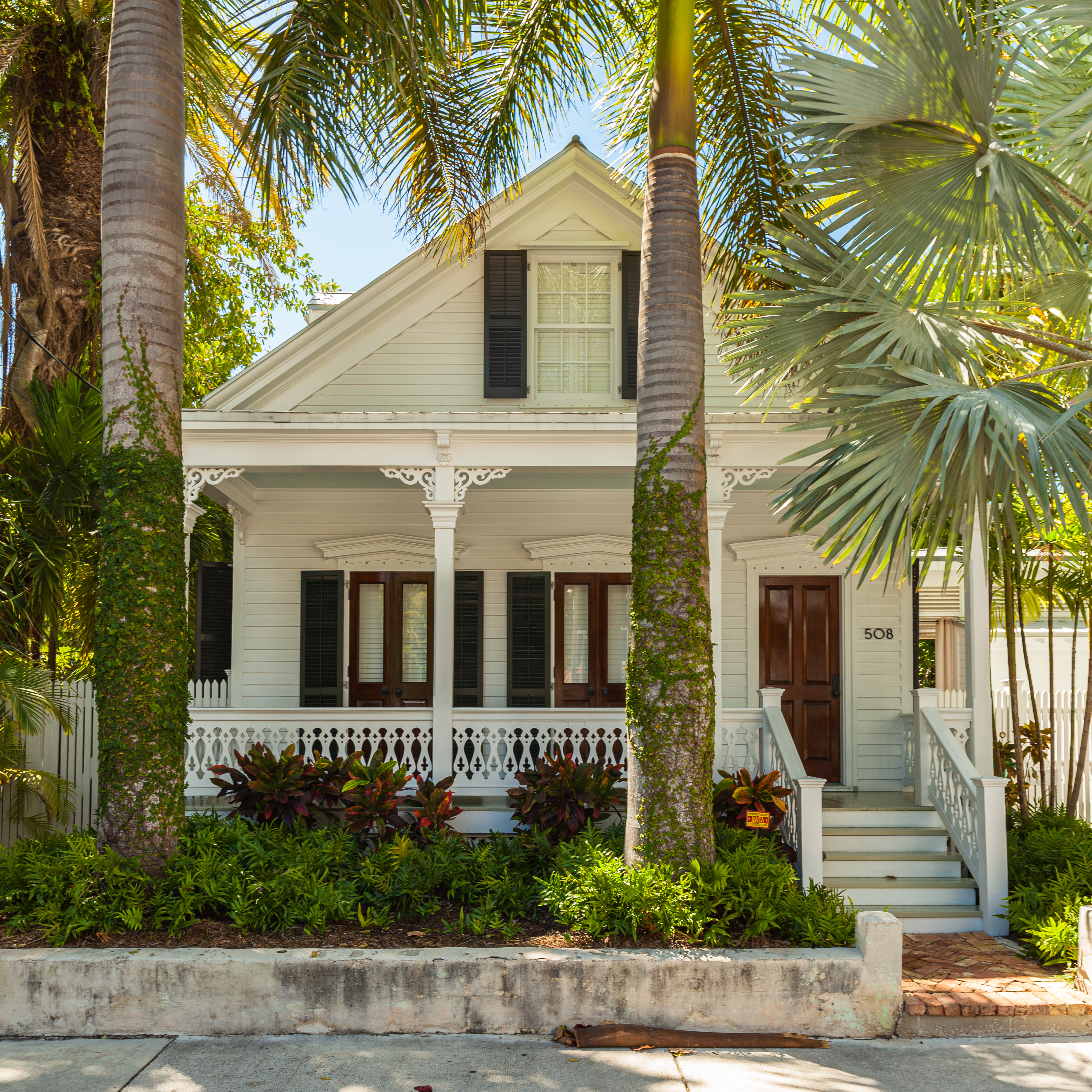 A charming, vintage white house with ornate trim, a wooden front door, and surrounded by lush tropical plants and palm trees. The house number 508 is visible