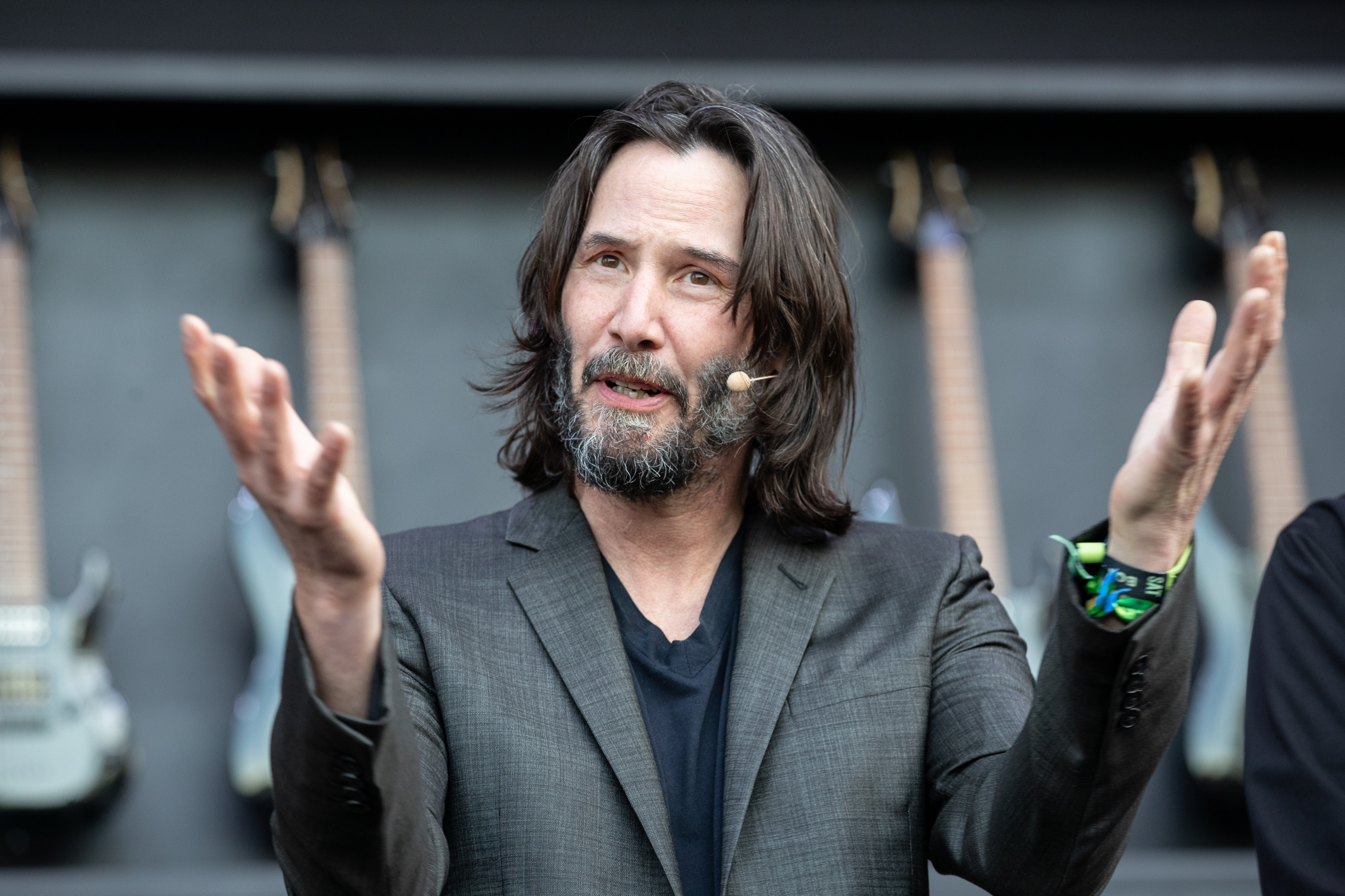 Keanu Reeves, with expressive hand gestures, wears a suit jacket and a microphone headset while speaking at an event with guitars in the background