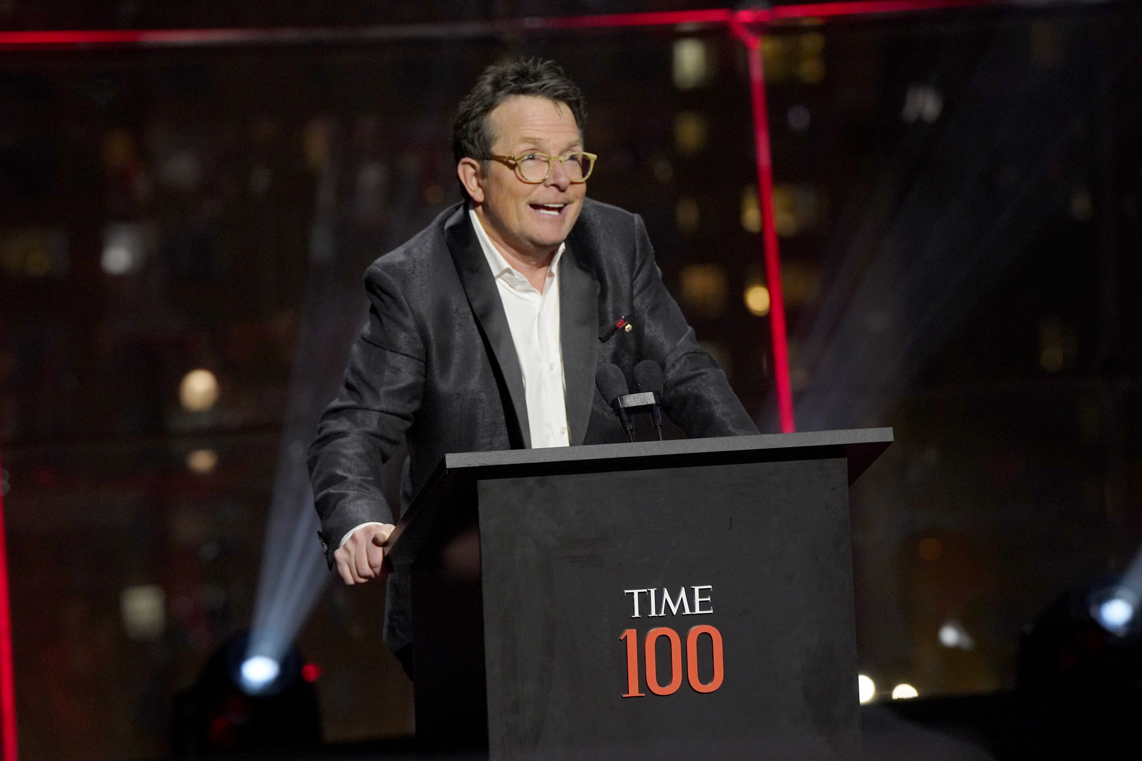 Michael J. Fox stands at a podium with "TIME 100" displayed, speaking at the event. He wears a dark suit jacket over a white shirt