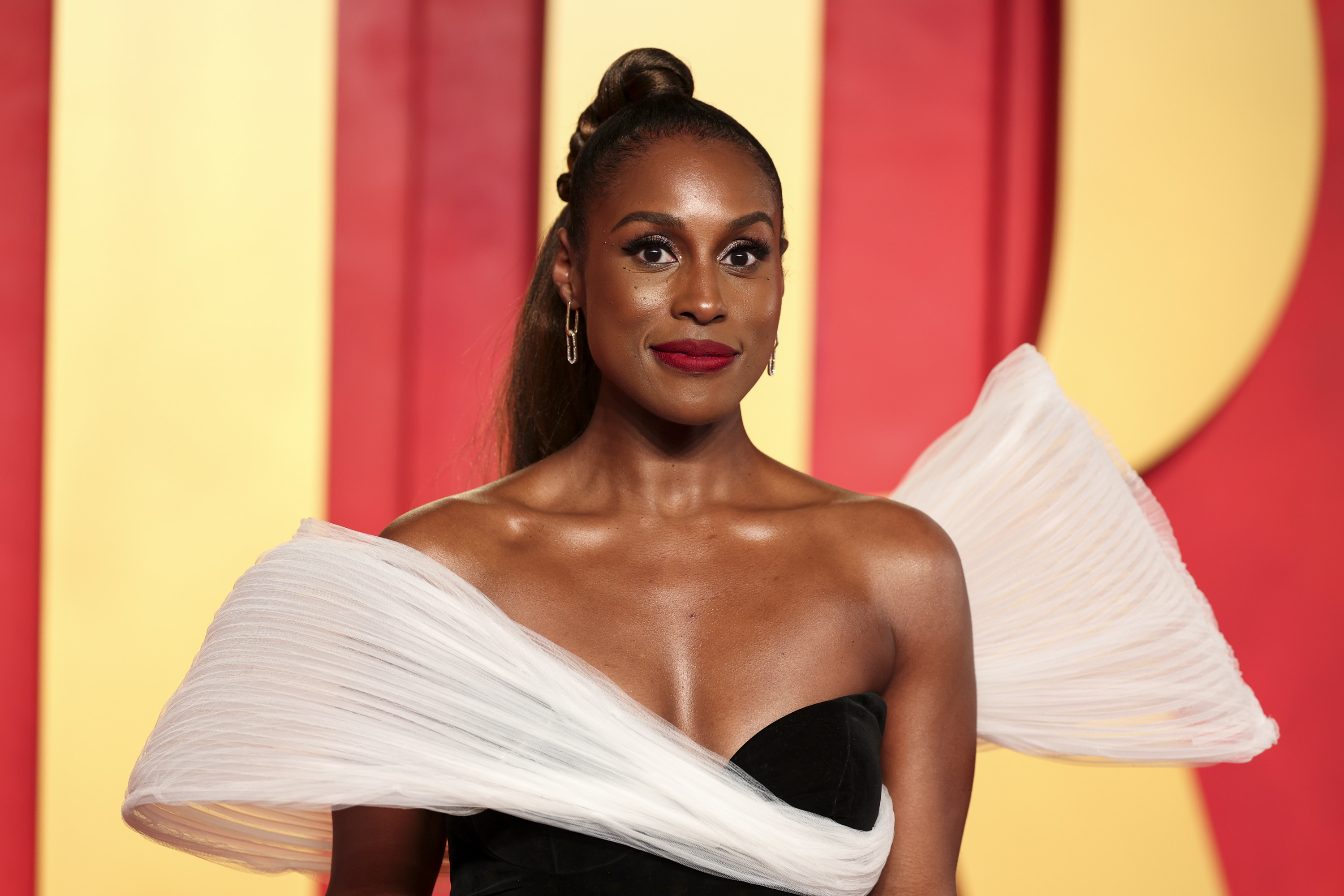Issa Rae on a red carpet wearing a strapless black dress with a unique white tulle detail across her chest and shoulders