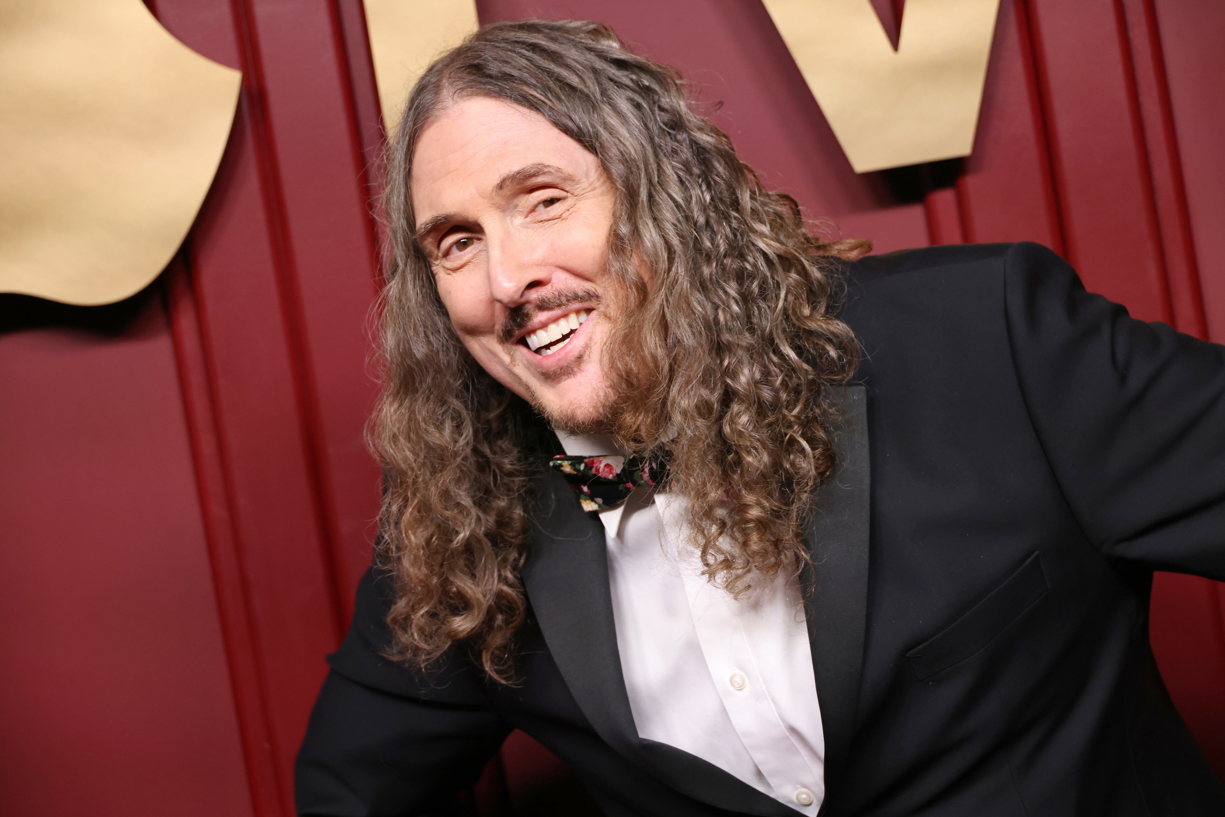 Weird Al Yankovic smiling, wearing a black tuxedo and a floral bow tie at a formal event