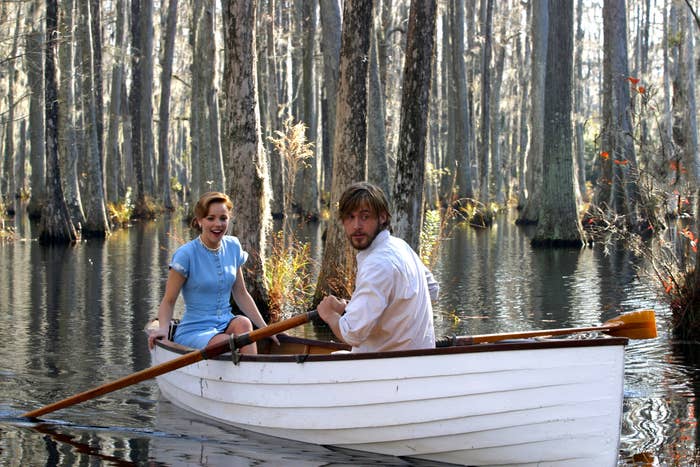 Rachel McAdams in a blue dress and Ryan Gosling in a white shirt sit in a boat on a lake surrounded by trees, in a scene from "The Notebook."