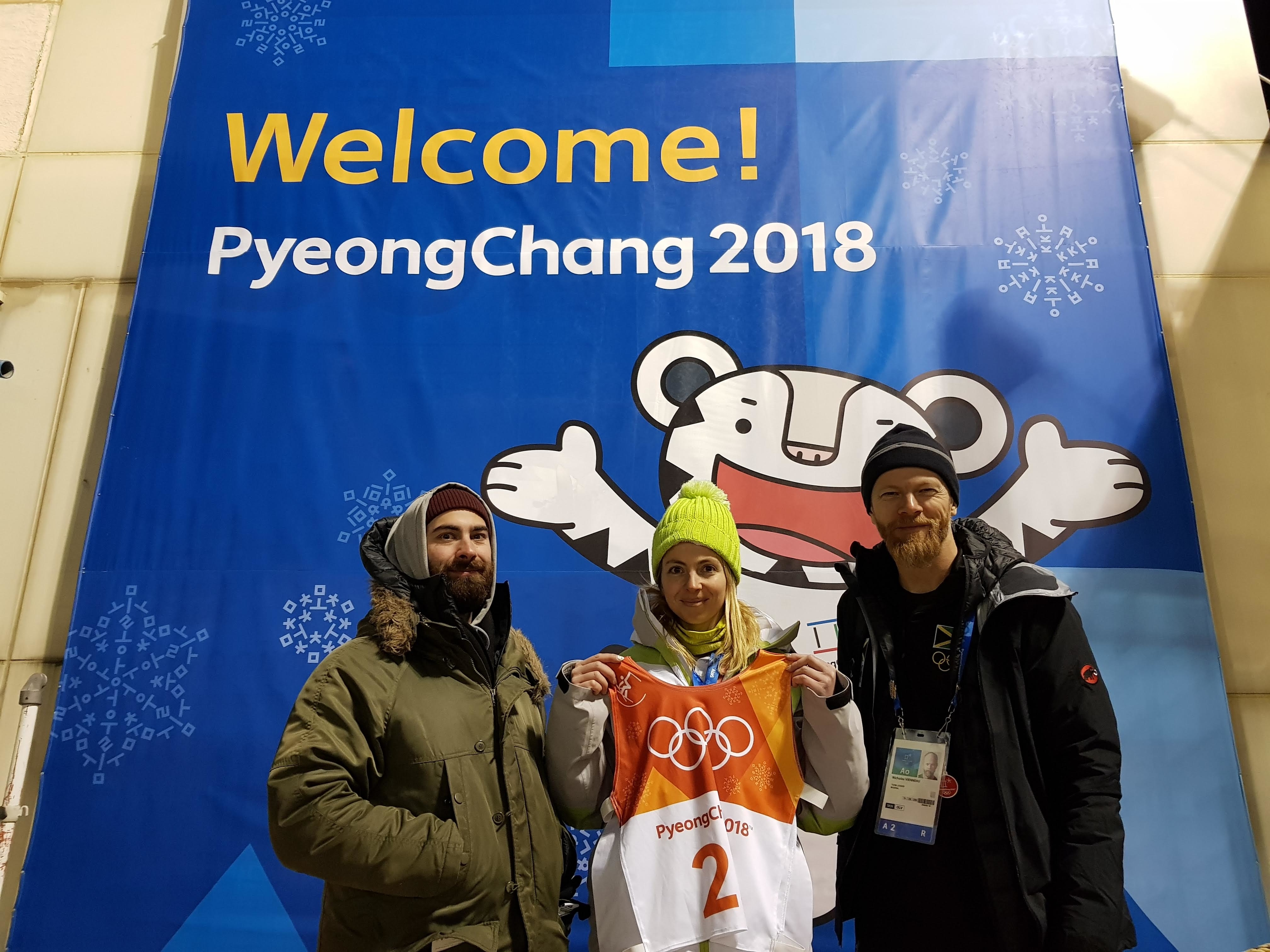 Allen Blackwell, Elizabeth Swaney, and Nicholas Vienneau, skeleton and new ski coaches, stand in front of a "Welcome! PyeongChang 2018" banner, with Elizabeth holding an Olympic bib