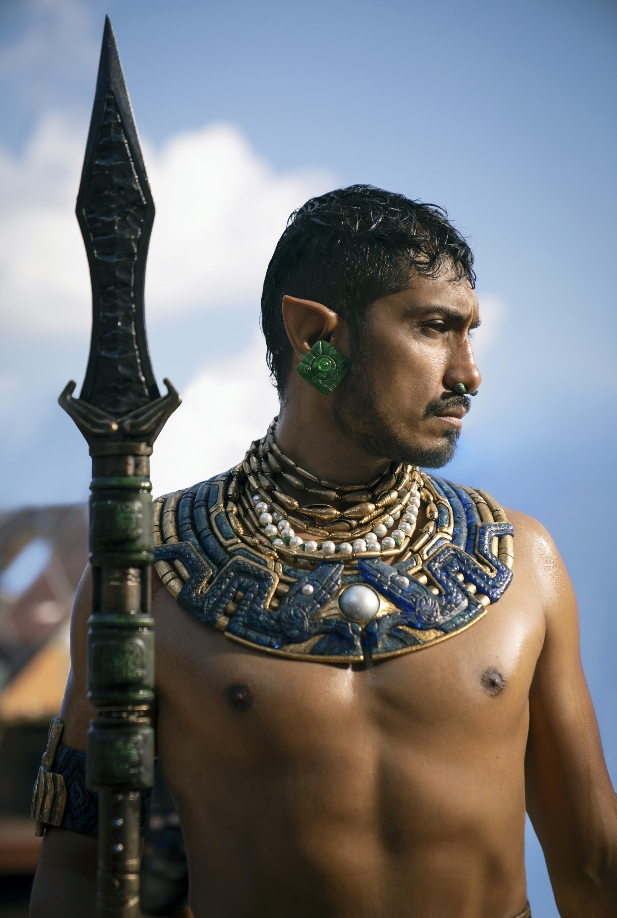 Tenoch Huerta wearing elaborate jewelry and holding a spear with a stern expression in a scene from "Black Panther: Wakanda Forever"