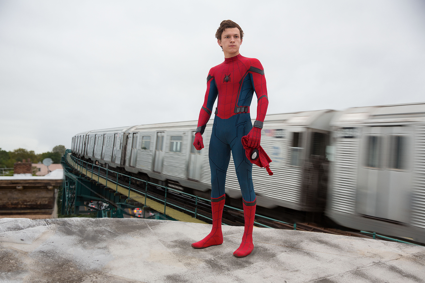 Tom Holland as Spider-Man stands on a rooftop, holding his mask, with a subway train passing in the background in a scene from "Spider-Man: Homecoming"