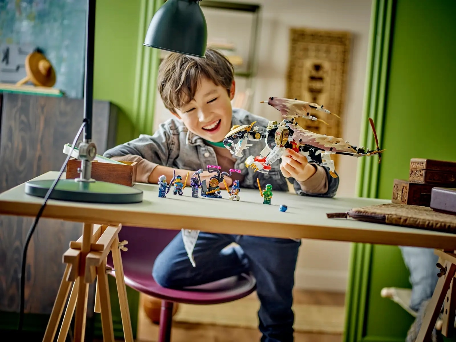 A boy smiles and plays with LEGO figurines and a dragon on a wooden table in a cozy room