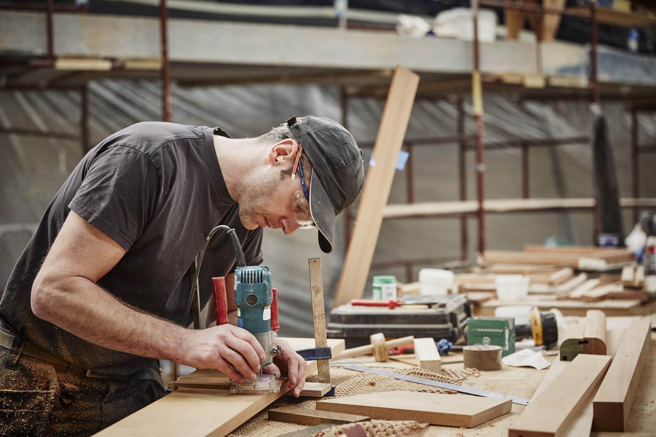 A craftsman in a workshop uses a power tool to shape wood. Safety glasses and ear protection are worn for safety. Various tools and materials are seen on the workbench