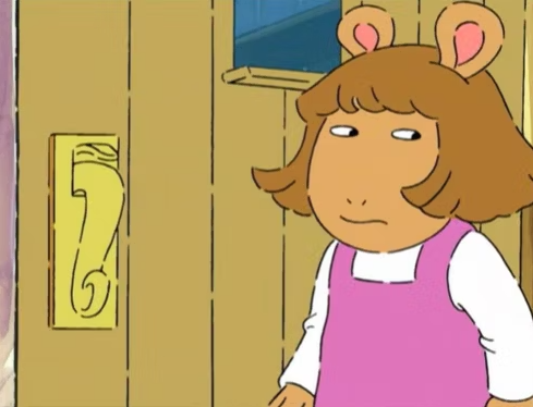 D.W. from Arthur stands near a door, wearing a pink dress over a white shirt, with a skeptical expression on her face