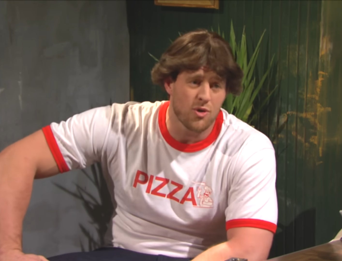 A man sitting at a table wearing a white t-shirt with the word "PIZZA" printed on it in red letters