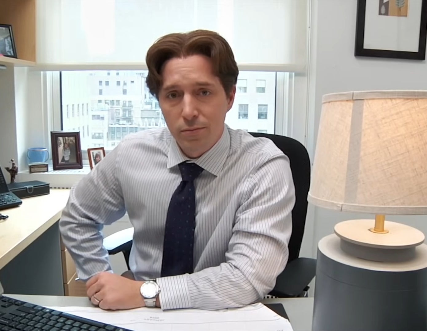 A man sits in an office at a desk with papers, a computer, and a lamp, looking directly at the camera