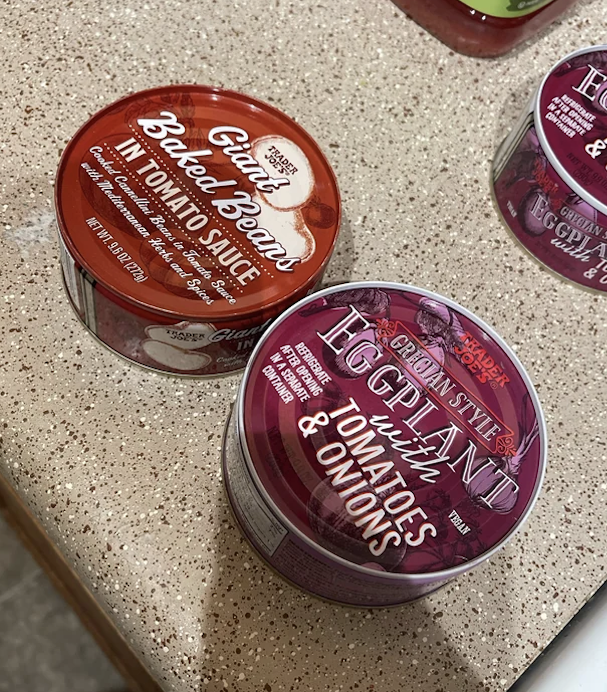 Two canned goods: one labeled &quot;Baked Beans in Tomato Sauce,&quot; the other &quot;Eggplant with Tomatoes &amp;amp; Onions.&quot; Both are placed on a countertop