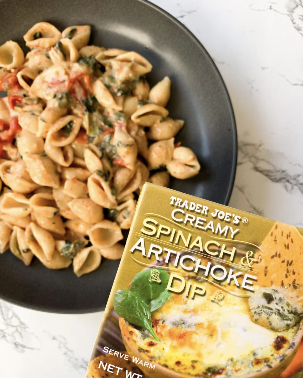 A plate of pasta with spinach and tomatoes next to a Trader Joe's Creamy Spinach &amp;amp; Artichoke Dip package
