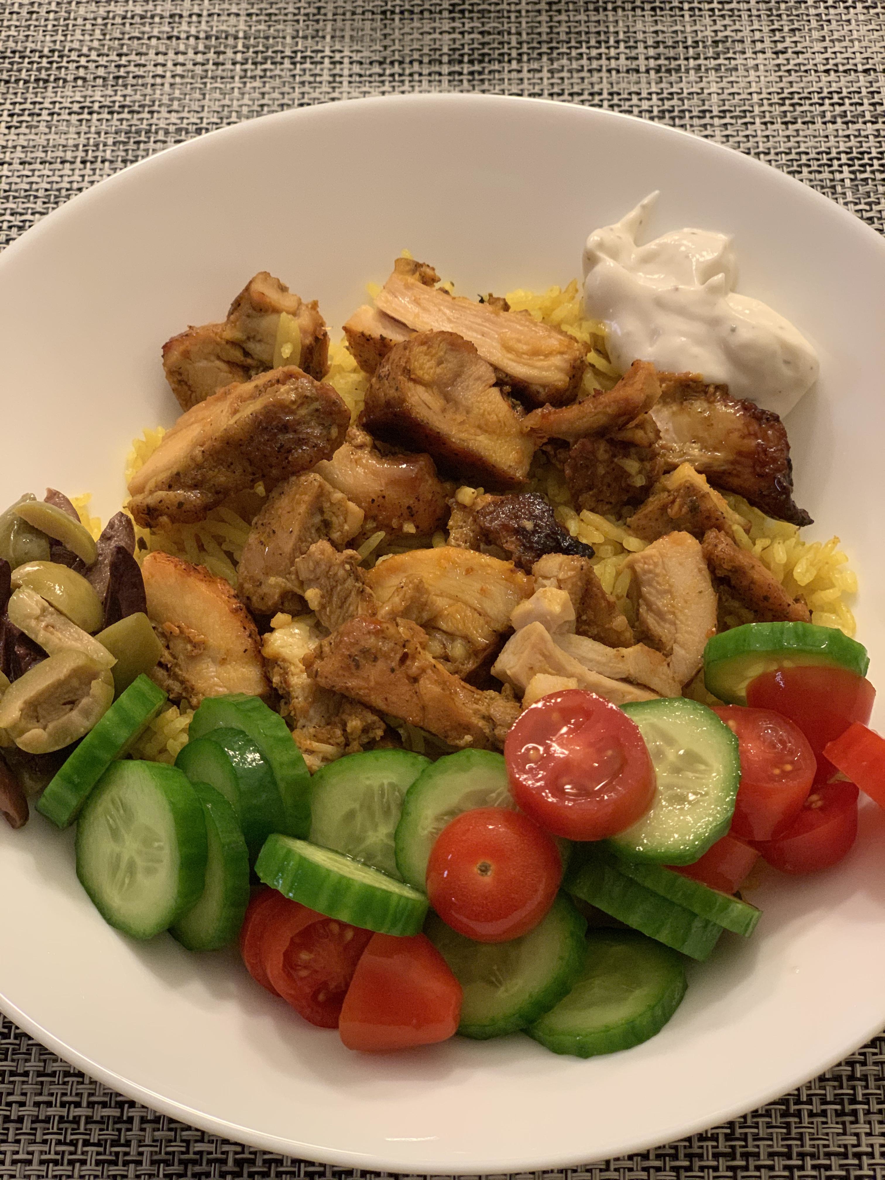 A bowl of seasoned chicken pieces on yellow rice, with sides of sliced cucumbers, cherry tomatoes, olives, and a dollop of sauce