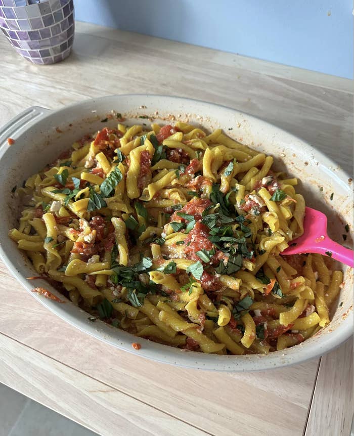 A dish of pasta topped with tomato sauce, green herbs, and cheese, served in a white dish with a pink spatula on the side