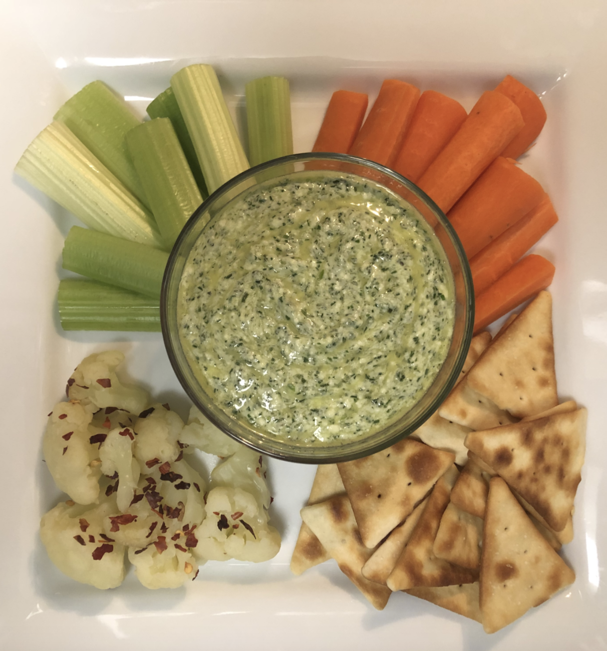 A plate of celery, carrot sticks, pita bread, and cauliflower with red pepper flakes surrounds a bowl of green vegetable dip