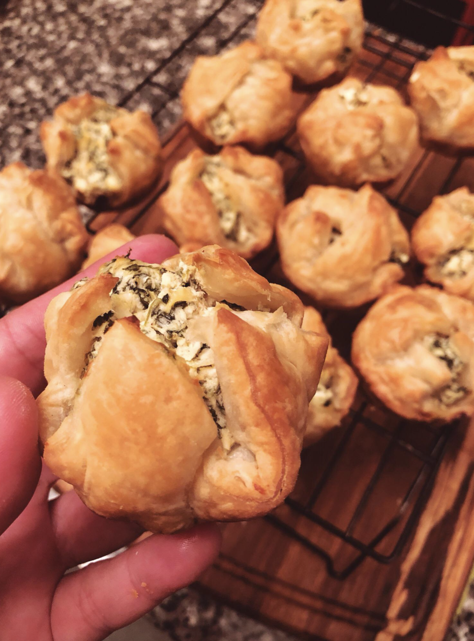 Hand holding a freshly baked puff pastry filled with spinach and cheese, with more pastries on a cooling rack in the background