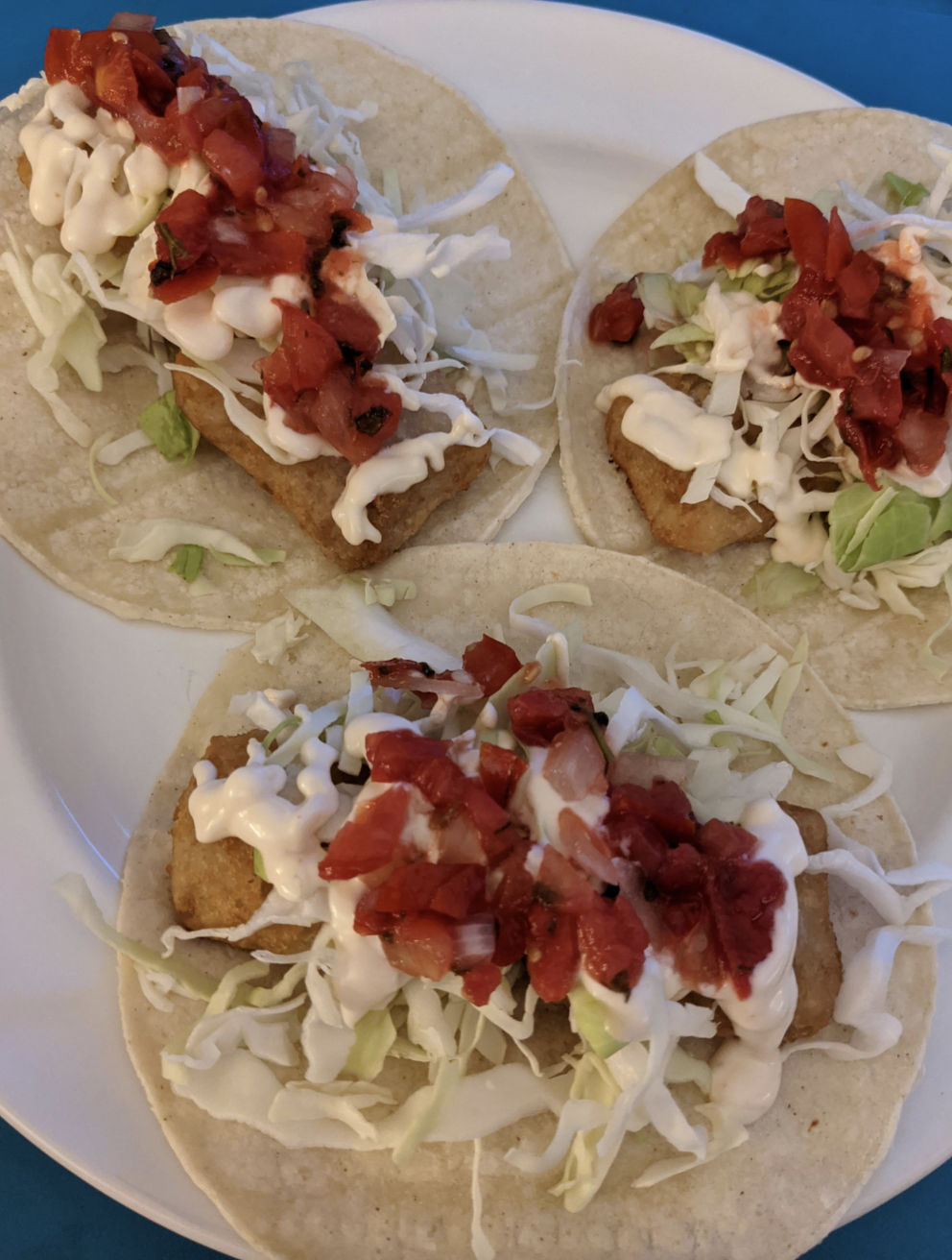 Three fish tacos with shredded cabbage, diced tomatoes, and a creamy white sauce on a white plate