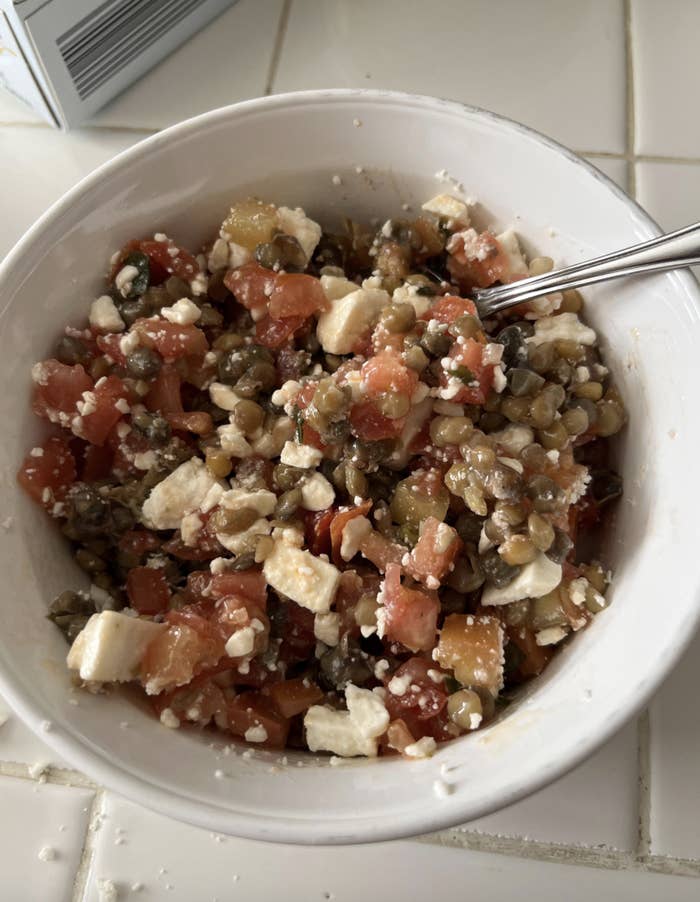 A bowl filled with a salad consisting of diced tomatoes, lentils, feta cheese crumbles, and chopped vegetables, with a metal spoon inside