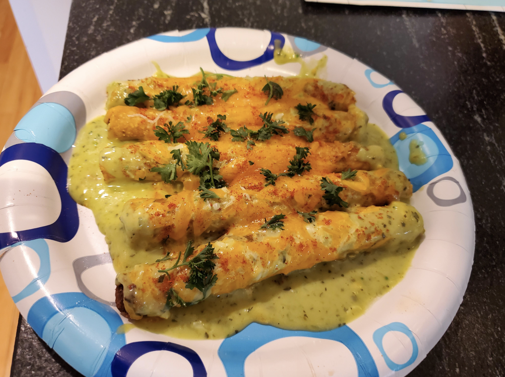 Five enchiladas covered in sauce and garnished with cilantro on a white paper plate with blue and green designs
